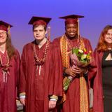 Temple Grandin School Photo - Group of graduates in caps and gowns celebrating on stage, one holding a bouquet.